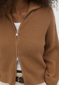 Bruine geribbelde gebreide cardigan met een zilveren ritsluiting. Kenmerkt zich door een hoge kraag en een ontspannen pasvorm, gedragen over een wit top.