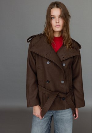 Une femme pose devant un fond gris de studio, vêtue d’un manteau marron croisé à ceinture par-dessus un haut rouge et un jean bleu.