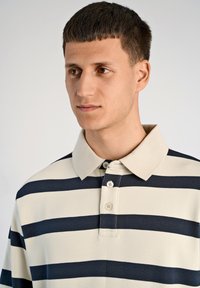 Polo w paski z beżowymi i granatowymi poziomymi pasami, zapięcie na guziki i klasyczny kołnierzyk, z gładką fakturą tkaniny.