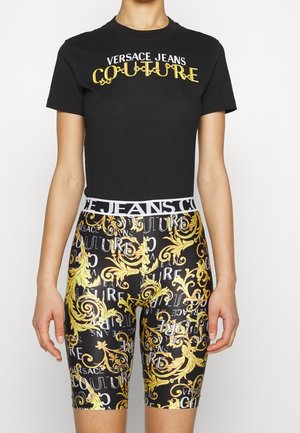 Kobieta nosząca czarny t-shirt Versace Jeans Couture oraz pasujące czarne szorty ze złotym wzorem barokowym i białym napisem logo.