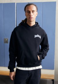 Black oversized hoodie with front pocket and ribbed cuffs. Features "BENEFIT" logo in white. Worn over a white shirt and dark pants.