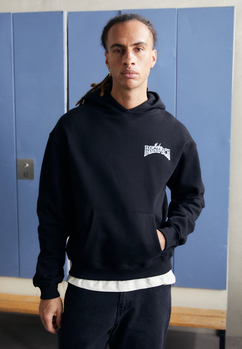Black oversized hoodie with front pocket and ribbed cuffs. Features "BENEFIT" logo in white. Worn over a white shirt and dark pants.