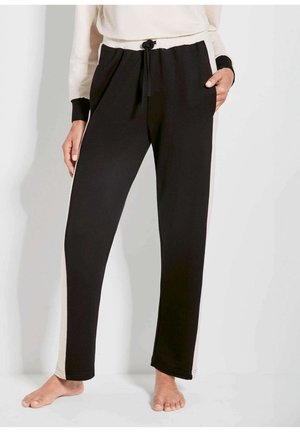 Persoon die zwarte en beige trekkoordenbroek met zijstrepen draagt, blootsvoets staand met één hand in de zak tegen een effen achtergrond.