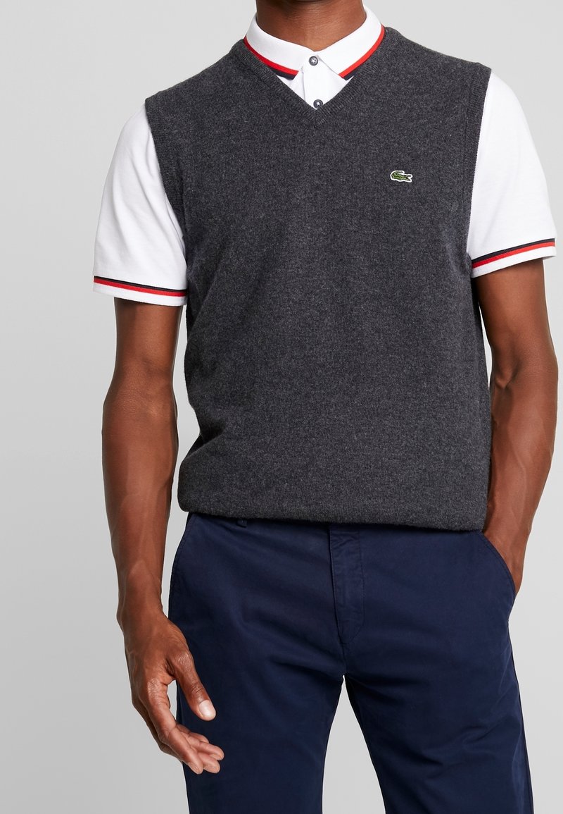 Grå stickad väst med V-ringning över en vit pikétröja med röda och svarta randiga detaljer, buren med marinblå byxor. Broderad logotypdetalj.