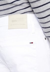 Vita denimbyxor med en läderetikett som har texten "HILFIGER DENIM." De har en liten tri-färgad logotyp på bakfickan och subtila sömnadsdetaljer.