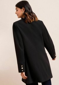 Černý kabát z vlněného materiálu s elegantním střihem, širokými rukávy a zlatými knoflíky na manžetách. Vyznačuje se čistou, strukturovanou siluetou.