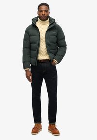 Mørkegrøn pufferjakke med hætte, cremefarvet kabelstriktrøje, mørke jeans og brune lædersko med lyse snørebånd. Enkelt, afslappet outfit.
