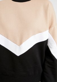 Sweatshirt coloré avec des panneaux beige, blanc et noir ; col noir côtelé ; texture douce ; design en chevrons obliques sur la poitrine.