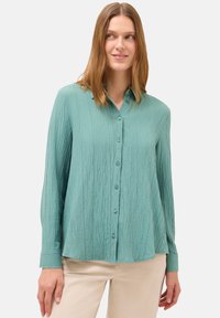 Camicia button-up di un azzurro chiaro con superficie testurizzata, maniche lunghe e orlo arrotondato. Presenta bottoni coordinati e colletto classico.
