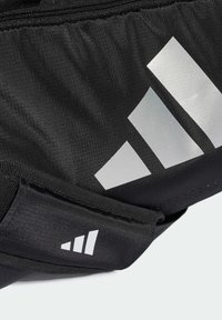 Svart stoffveske med sølvfarget Adidas-logo. Har glidelås og teksturert overflate. Pyntet med tre striper mønster og justerbar stropp.