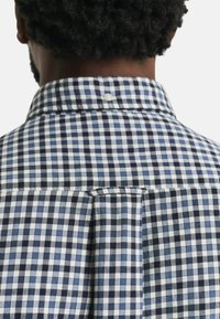 Camisa de cuadros con un patrón a rayas azules y blancas, cuello abotonado y un pliegue en la parte posterior. Tejido de algodón de textura suave.