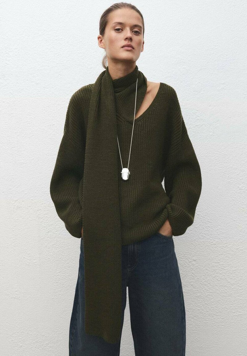 Maglione verde scuro lavorato a maglia con taglio oversize e scollo ampio, abbinato a una sciarpa coordinata. La modella indossa jeans e una collana d'argento.