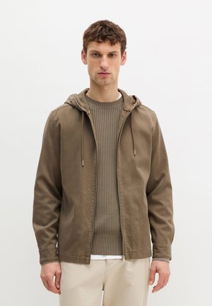 Jonge man die een bruine zip-up capuchontrui draagt over een geribbelde trui en beige broek, staand tegen een effen witte achtergrond.