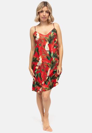 Vrouw draagt een rode mouwloze jurk met print van kaketoe en tropische bladeren, staat blootsvoets tegen een effen lichte achtergrond.