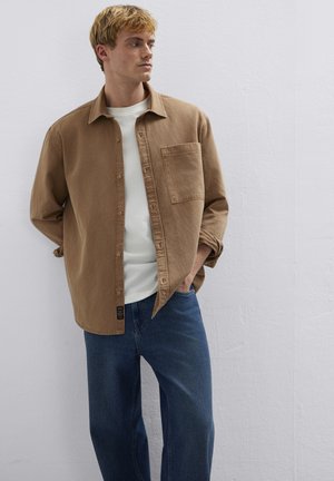 Junger Mann, der ein braunes Hemd über einem weißen T-Shirt und einer blauen Jeans trägt, mit den Händen in den Taschen stehend und seitlich blickend.