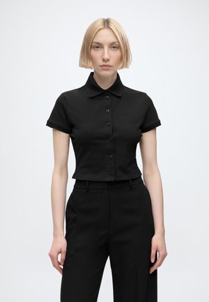 Jeune femme aux cheveux blonds courts portant une chemise courte noire boutonnée à manches courtes et un pantalon noir sur un fond uni.