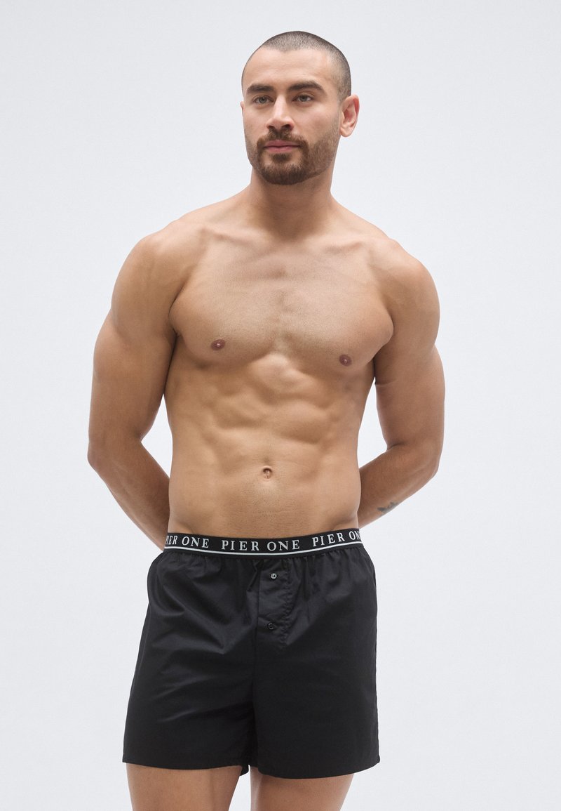 Shorts de bain noirs avec une ceinture élastique ornée d'un logo. Les shorts ont une fermeture boutonnée et une coupe décontractée. Matière lisse.