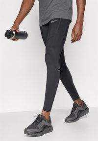 Μαύρα αθλητικά κολάν με στενή εφαρμογή και διακριτές ραφές, συνδυασμένα με σκούρα αθλητικά παπούτσια, κρατούμενα από ένα άτομο που κρατάει ένα μπουκάλι νερού.