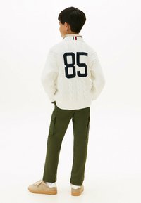 Hvit kabelstrikket genser med marineblått tall "85" på ryggen, sammen med olivengrønne cargo-bukser og beige joggesko. Hvit bakgrunn.
