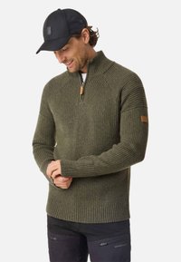 Olive green, ribbed-knit sweater with a quarter zipper and reinforced seams. Features a leather tag on the left sleeve. Worn with black cap.