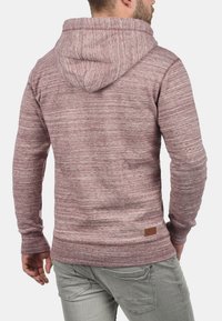 Bluza z kapturem w kolorze maroon i szarym, wykonana z teksturowanej tkaniny, z dużym kapturem i małą metką marki na dolnej części pleców. Cienka sylwetka.
