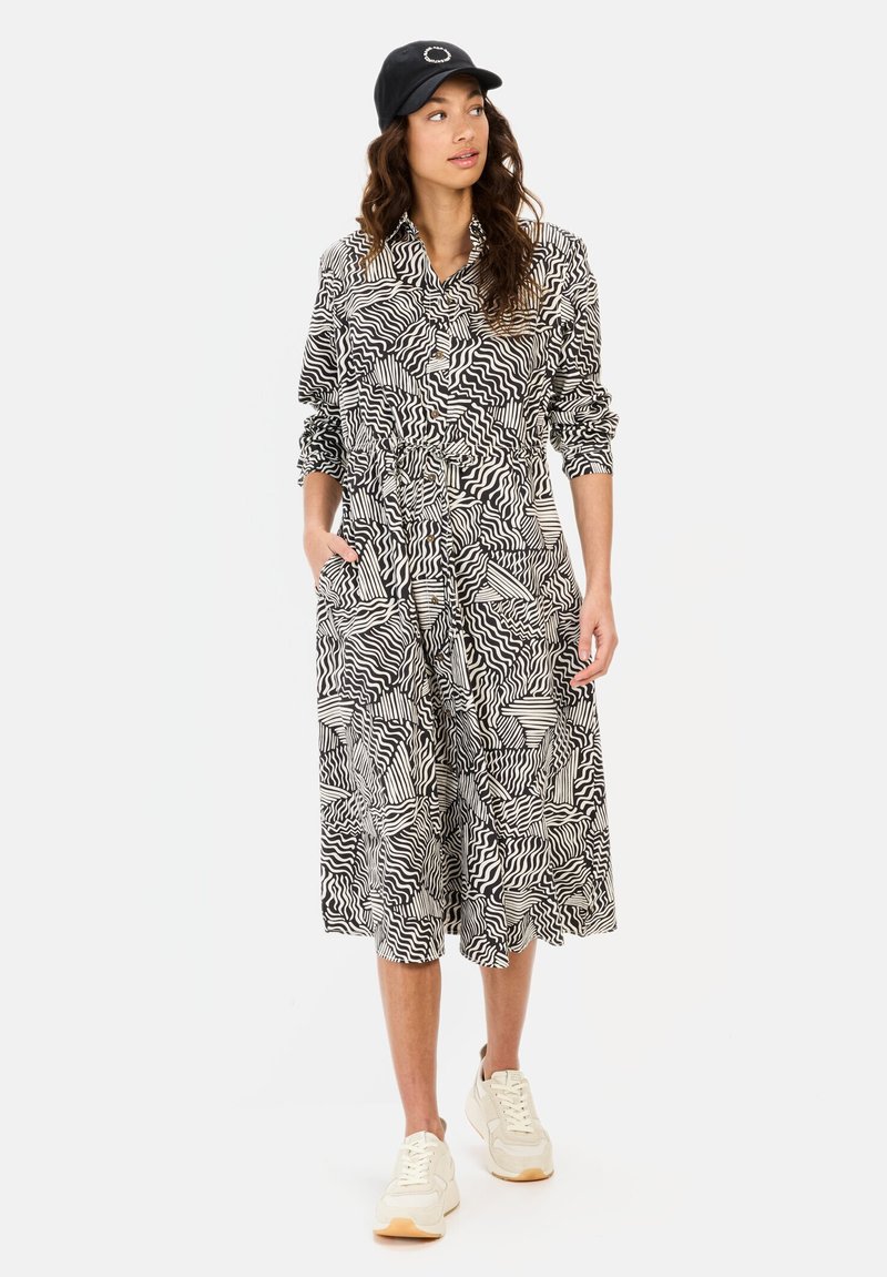 Schwarz-weiß gemustertes Kleid mit langen Ärmeln, durchgehender Knopfleiste und Taschen; kombiniert mit einer schwarzen Mütze und beigen Sneakers.