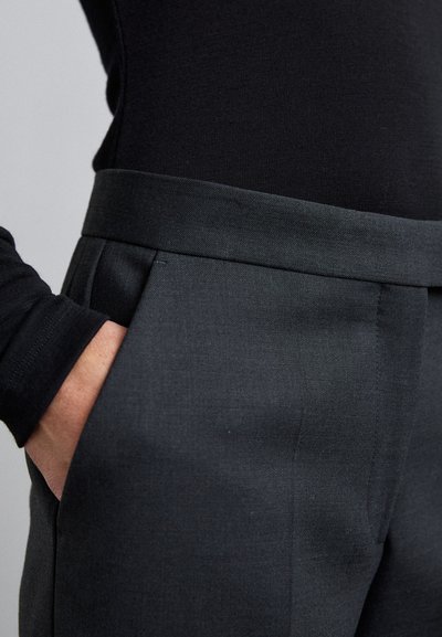 Person wearing high-waisted dark gray trousers with side pockets and a black long-sleeve top, hand casually in pocket.