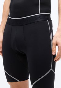 Άνδρας που φοράει μαύρα συμπιεστικά σορτς Under Armour με άσπρες ραφές και branded ζώνη μέσης, με το χέρι ακουμπισμένο στο μηρό.