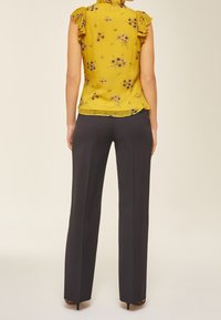 Femme debout, vue de dos, portant un chemisier sans manches jaune à motif floral et un pantalon noir à jambes larges avec des escarpins noirs.