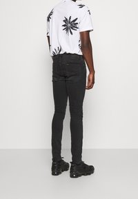 Man med svarta slim-fit jeans, svarta sneakers och en vit kortärmad skjorta med stora svarta blommönster, vänd bort.