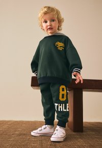 Dark green sweatshirt with yellow ‘Athletic Track & Field 1958’ print, paired with matching joggers featuring white lettering and number 88. White shoes.