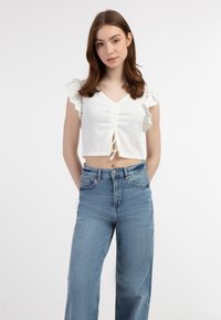 Hvid crop top med flæseærmer og snøre detalje foran, parret med højtaljede blå denim jeans. Glat stofstruktur.