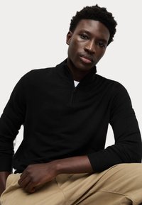 Sudadera negra con cremallera y cuello alto, con mangas ajustadas y una textura suave. Combinada con pantalones beige, el enfoque está en el diseño de la prenda.