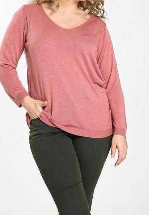 Kvinde iført en langærmet støvet pink sweater og mørkegrønne bukser, poserer med den ene hånd i lommen mod en ensfarvet baggrund.