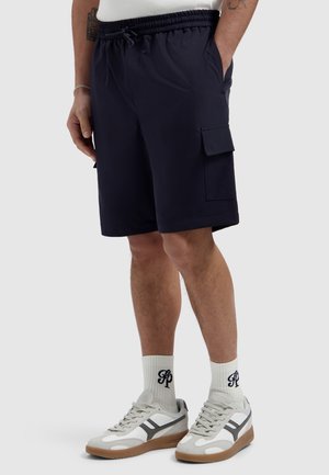 Mann trägt schwarze Cargoshorts mit elastischem Bund, weiße Socken mit schwarzem Logo und weiße und graue Turnschuhe, steht mit den Händen in den Taschen.