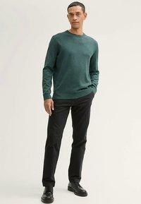 Donkergroene gebreide trui met een ronde hals, gecombineerd met zwarte broek en zwarte schoenen. De stof lijkt zacht met een gladde textuur.