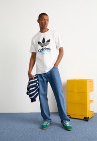 Maglietta bianca in cotone con logo Adidas nero e dettagli blu. Jeans blu, sneakers verdi con lacci bianchi. Maglione a righe blu scuro e blu. Unità di stoccaggio gialla.