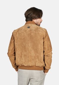 Chaqueta bomber de ante color topo con cuello y puños acanalados. Presenta un parche de logo cuadrado en la parte posterior y una superficie texturizada.