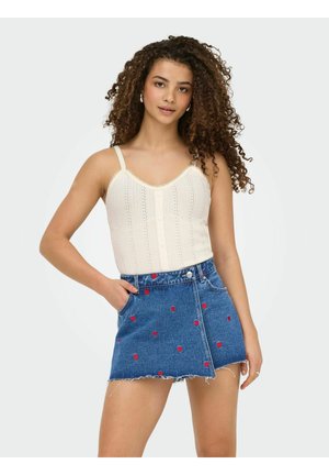 Žena kovrčave kose u krem pletenom topu i plavoj traper mini suknji s crvenim uzorcima srca, pozira s jednom rukom u džepu.