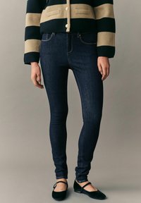 Jeans skinny de mezclilla azul oscuro con un corte ajustado y detalles de costura sutiles, combinados con zapatos negros planos, sobre un fondo neutro.