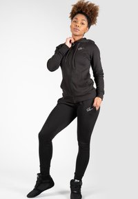 Zwarte rits-hoodie en bijpassende leggings, gemaakt van zacht materiaal. De hoodie heeft trekkoorden en een subtiel logo; de leggings hebben een zijzak.