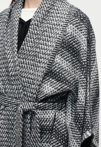 Παλτό με ασπρόμαυρο σχέδιο houndstooth, φαρδιές πέτρες, ζώνη δέσιμο στο μπροστινό μέρος και τσέπη στη δεξιά πλευρά, φορεμένο πάνω από ασπρόμαυρα τοπ.