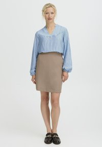 Mujer de pie vestida con una blusa de manga larga a rayas azul claro, una falda beige hasta la rodilla y mocasines negros, delante de un fondo blanco liso.