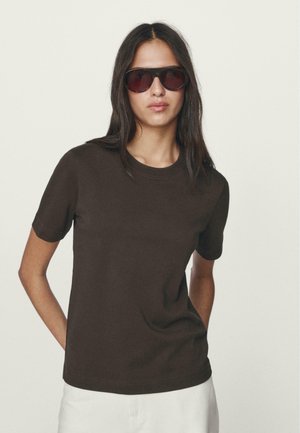 Brown cotton t-shirt with short sleeves, crew neck, and relaxed fit; worn with dark sunglasses against a neutral background.