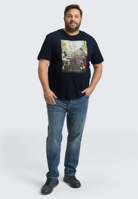 Navyblauw T-shirt met korte mouwen en een centrale grafische print van voertuigen in een bosachtige omgeving. Gekleed met lichtblauwe jeans en zwarte schoenen.