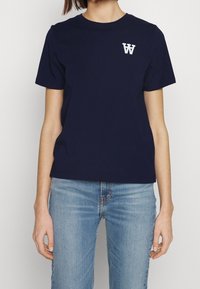 Námořnické modré bavlněné tričko s krátkým rukávem, kulatým výstřihem a bílým logem na hrudi. V kombinaci s světle modrými džíny.