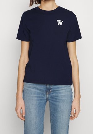 Camiseta de algodón azul marino de manga corta, con cuello redondo y un logo blanco en el pecho. Combinada con unos vaqueros de mezclilla azul claro.