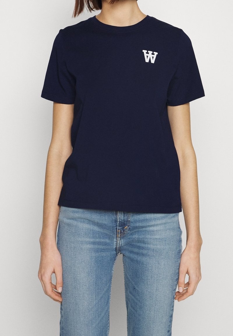 Námořnické modré bavlněné tričko s krátkým rukávem, kulatým výstřihem a bílým logem na hrudi. V kombinaci s světle modrými džíny.