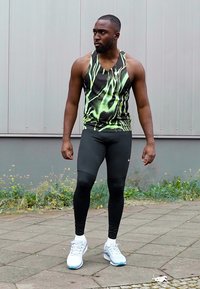 Canotta atletica nera e verde neon con un motivo fluido, abbinata a leggings neri aderenti e scarpe da ginnastica bianche.