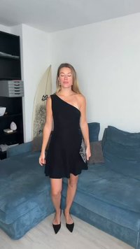 Crna haljina na jedno rame s prostranom suknjom; glatki materijal; uparena s torbicom teksture i crnim štiklama šiljastih vrhova. Jednostavan dizajn.